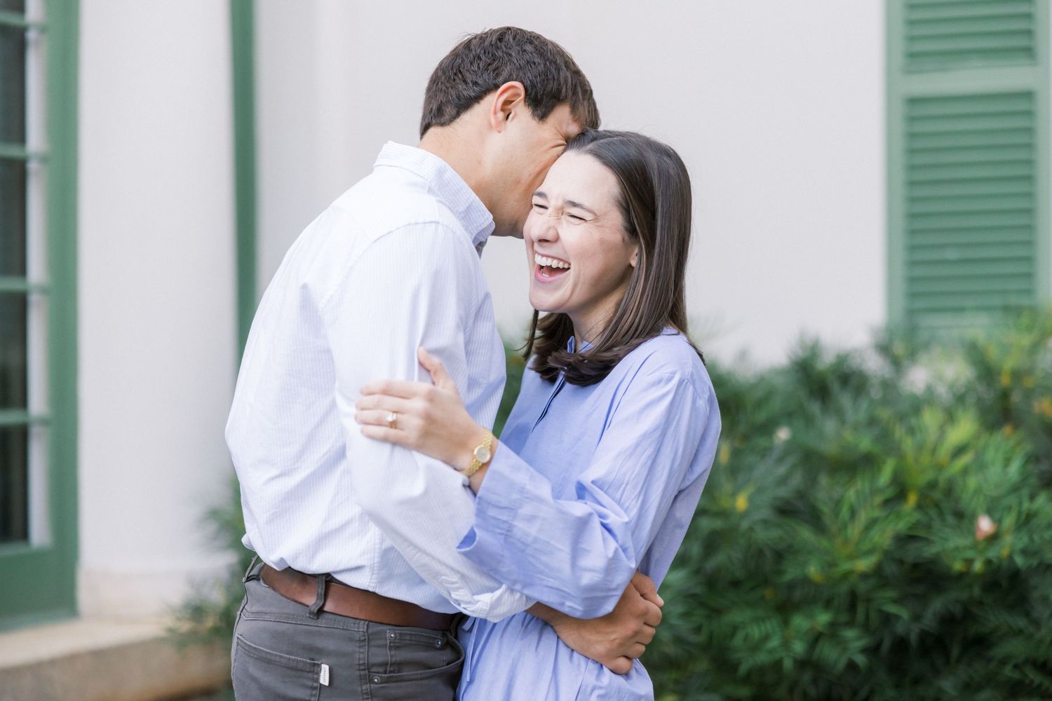 A couple laughs together while embracing outdoors near a building with green shutters and plants.