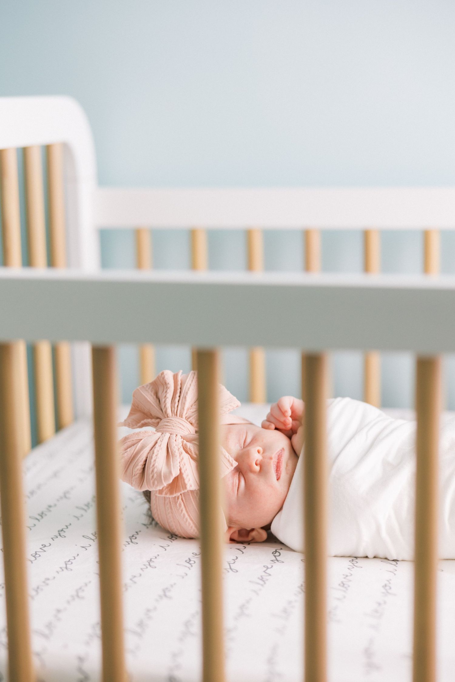 Sleeping newborn with pink bow lying peacefully in white wooden crib.