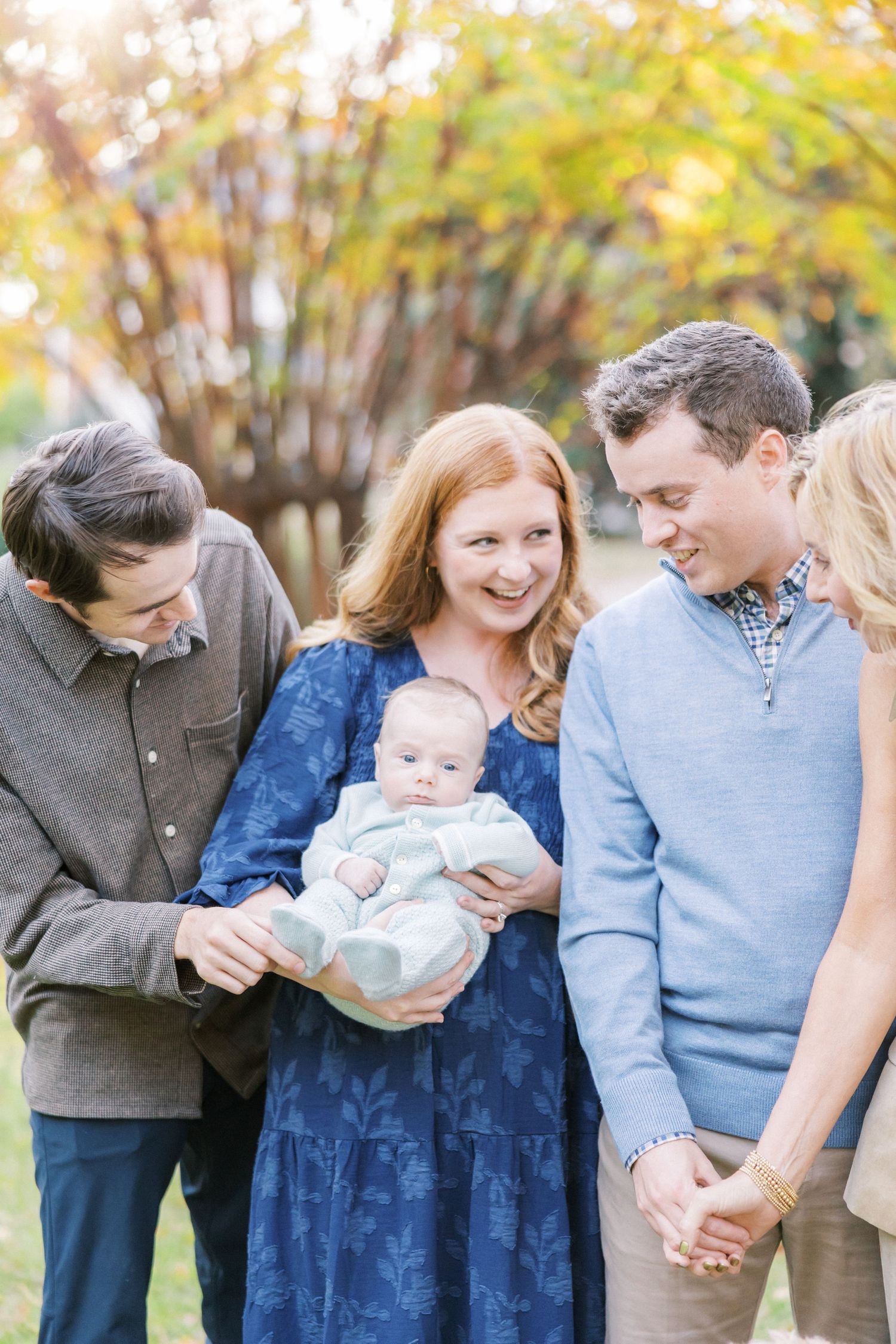 A small group gathered around a baby outdoors with fall foliage in the background.
