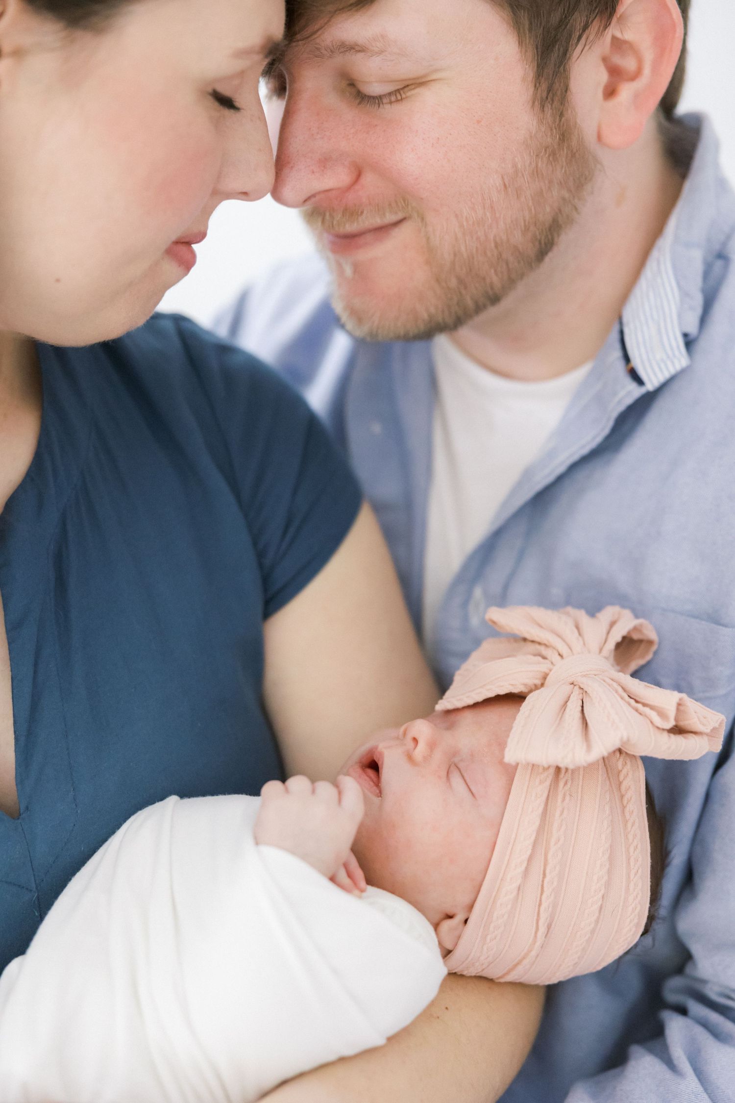 Parents lovingly gazing at their sleeping newborn baby wearing a pink bow headband.