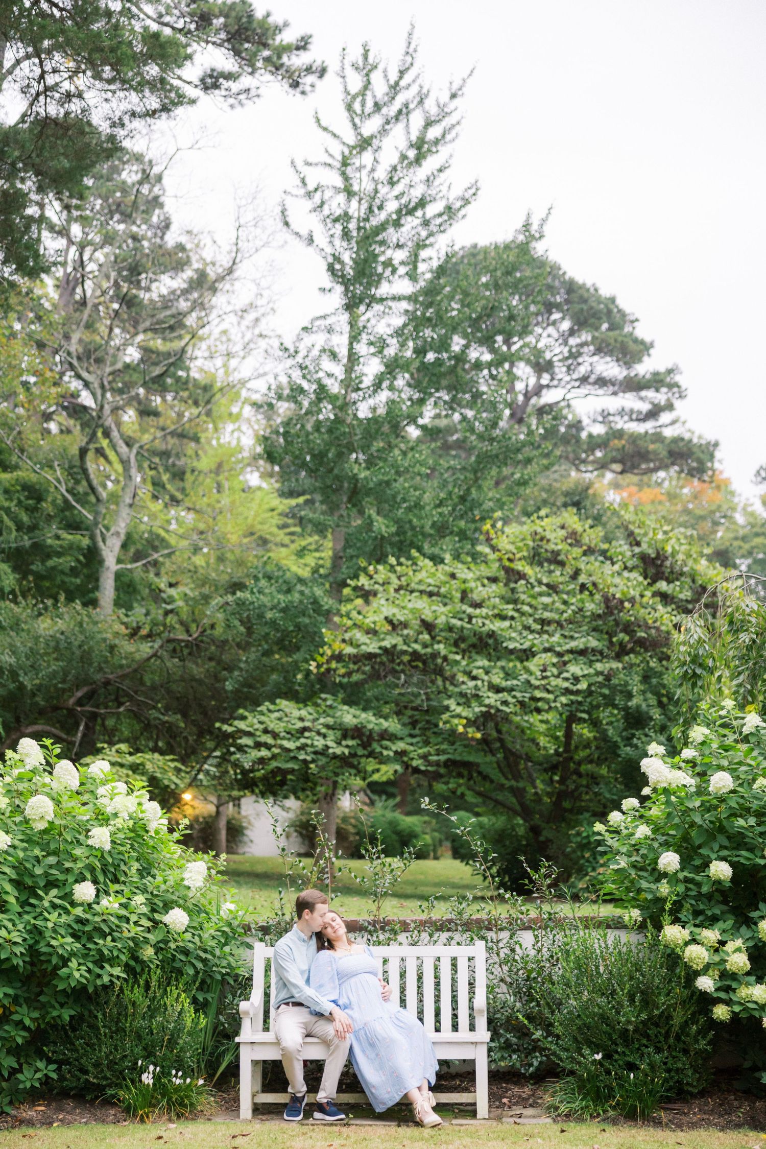 A couple shares a romantic moment on a white garden bench surrounded by lush greenery and hydrangea bushes.