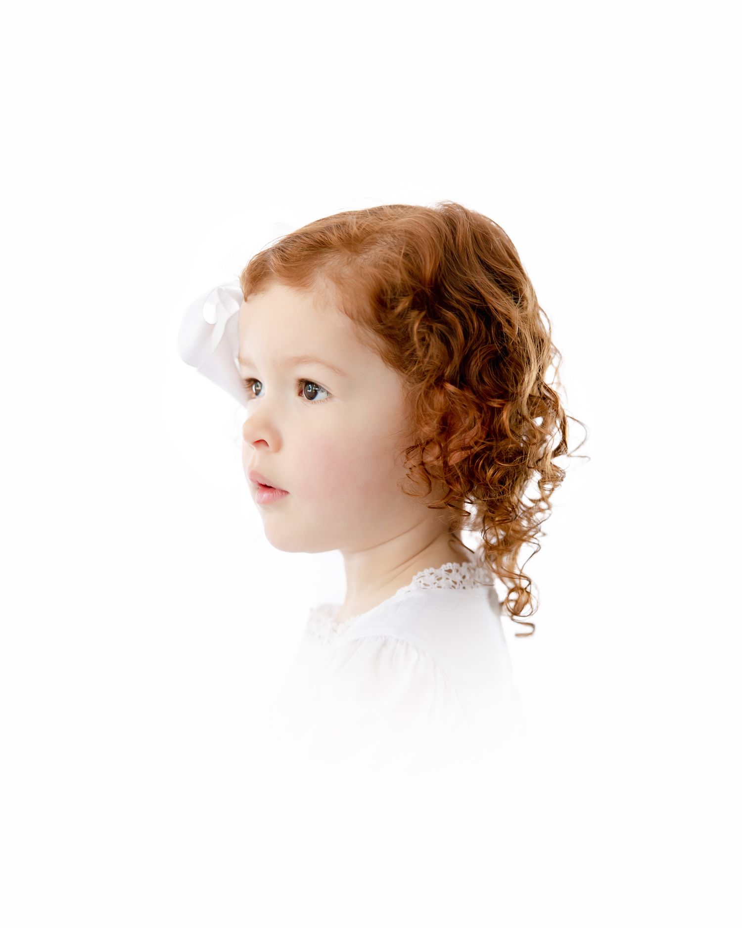 Profile view of a young child with auburn curly hair wearing a white top against a soft white background.