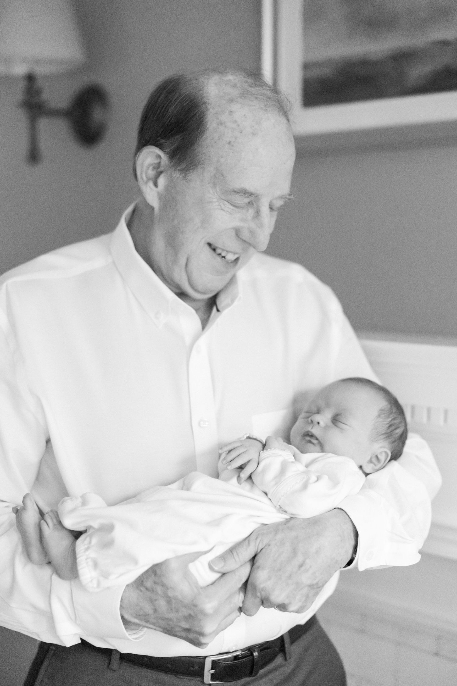 A joyful black and white portrait of a gentleman in white shirt holding and smiling at a sleeping newborn.