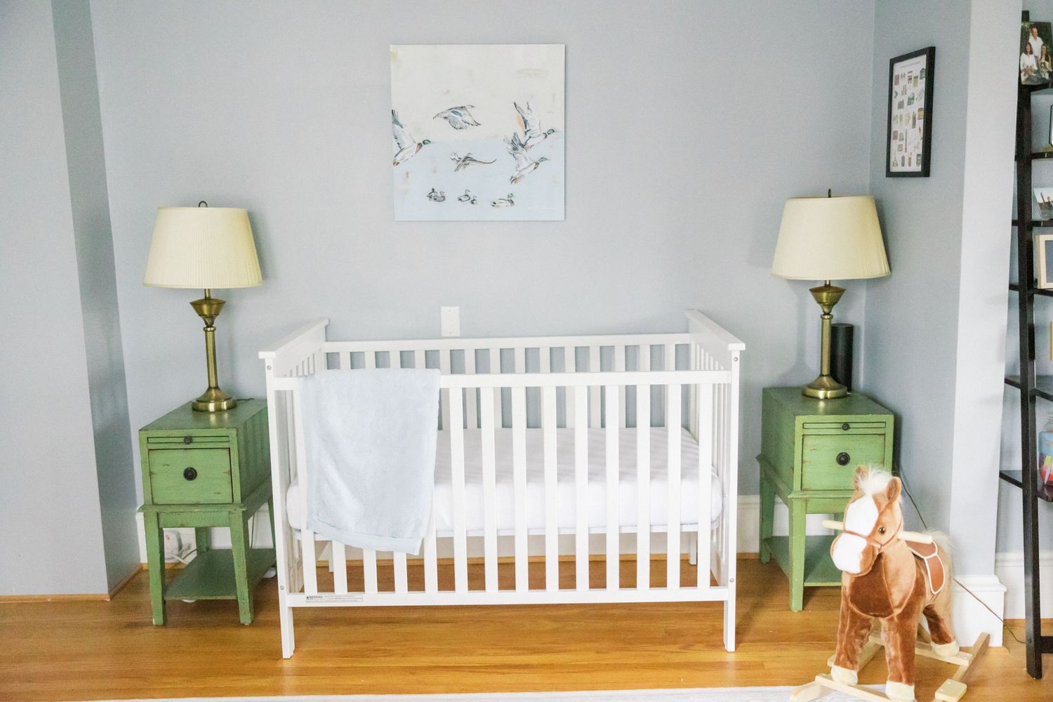 Minimalist nursery room with white crib, matching green side tables, bird artwork and stuffed animal decor.