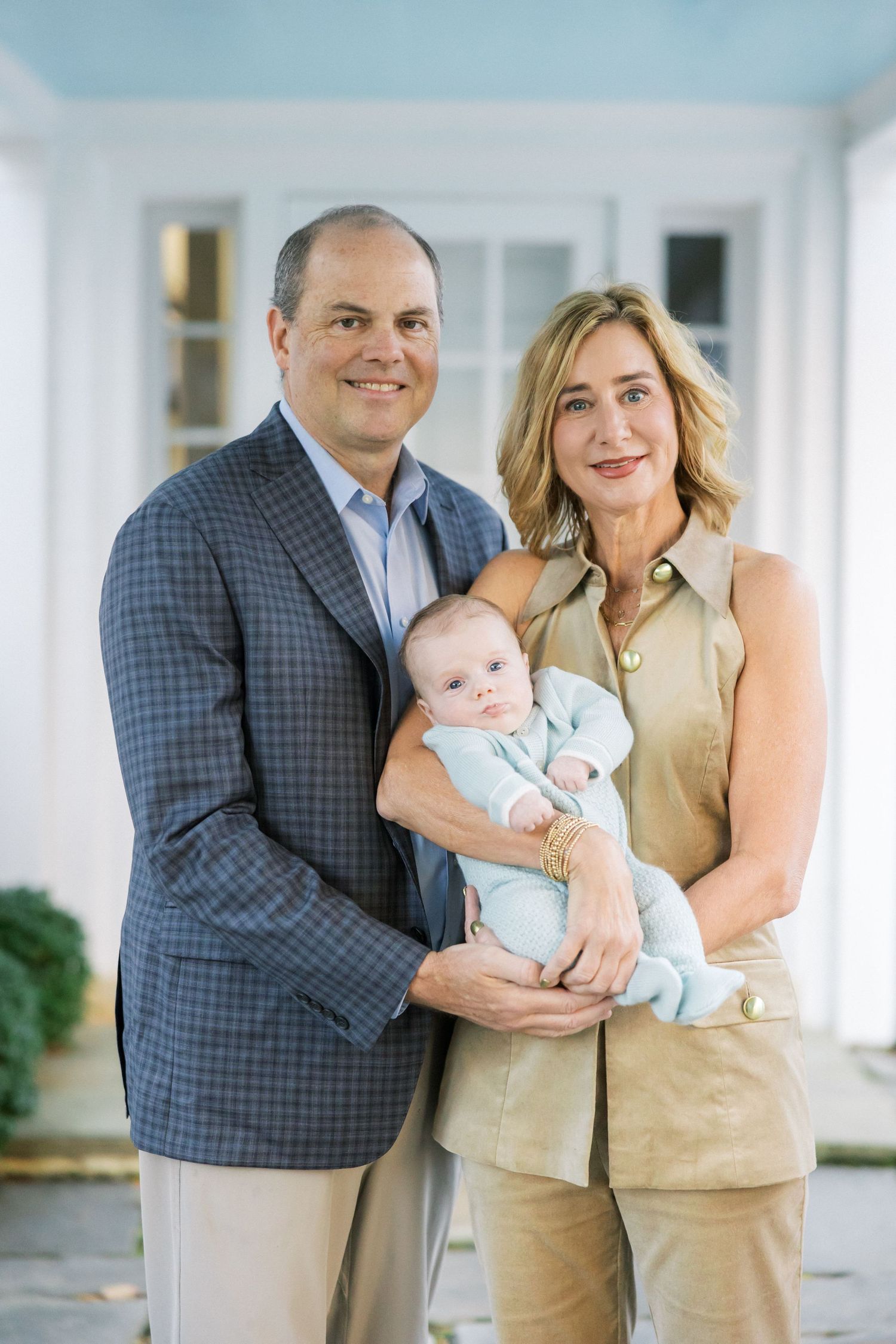A couple in business attire holds an infant in a light blue outfit while posing by a white column.