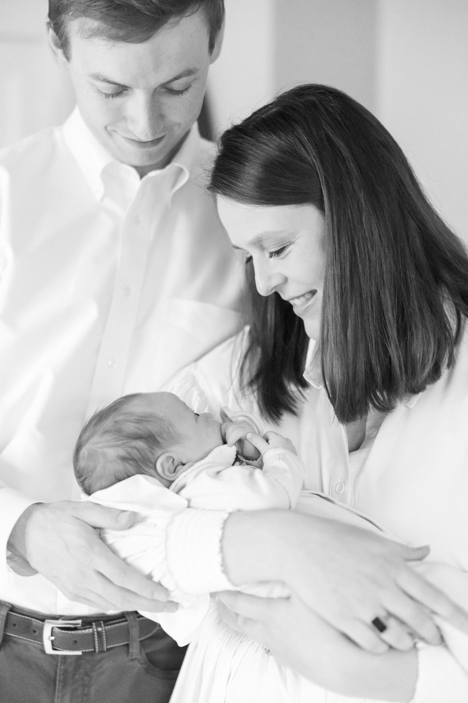 A black and white portrait of new parents tenderly holding their sleeping newborn baby swaddled in white.