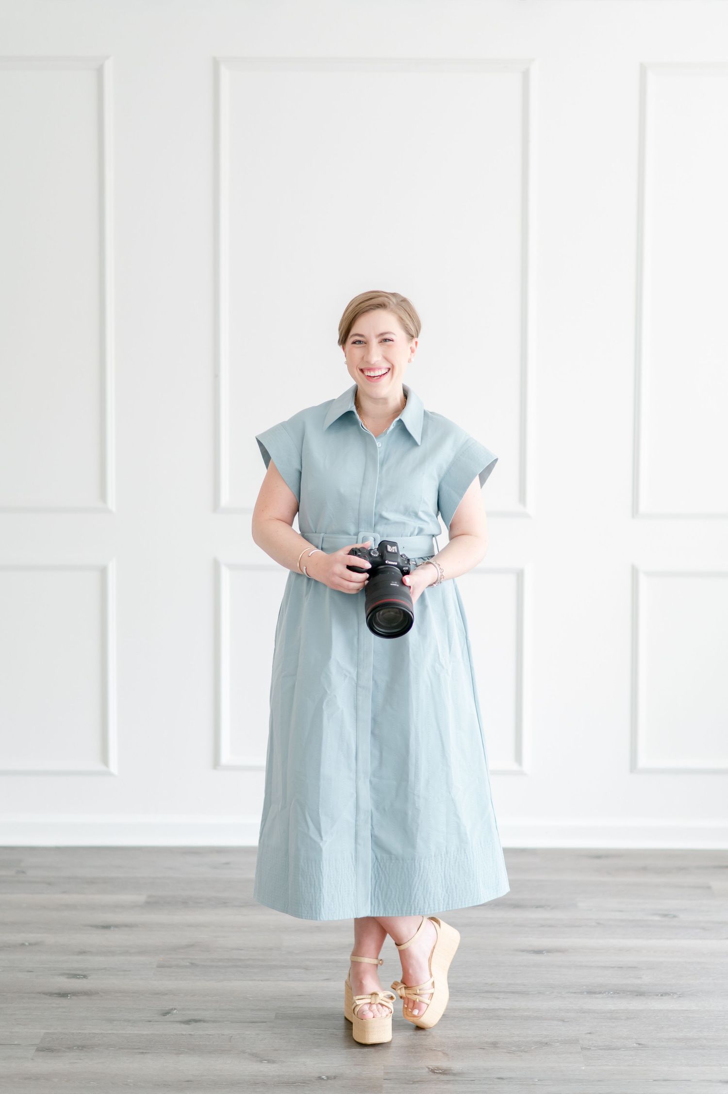 Professional photographer in light blue dress holding camera against white wall backdrop.