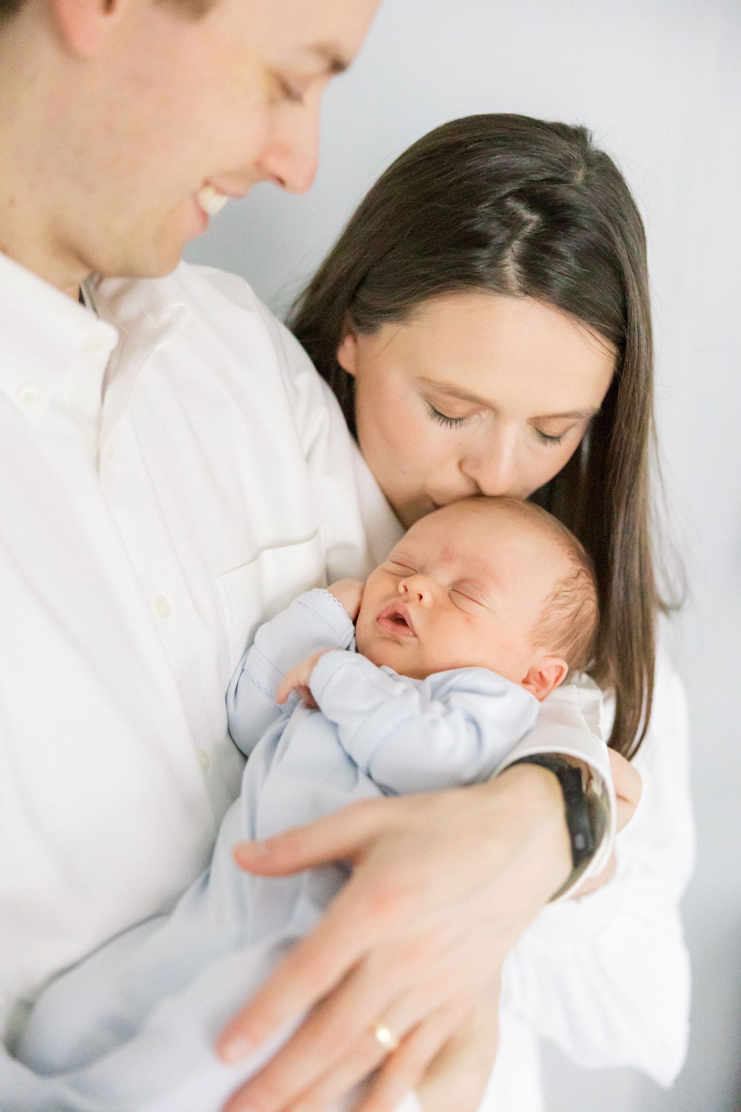 Parents in white shirts share a tender moment with their sleeping newborn baby wearing light blue.