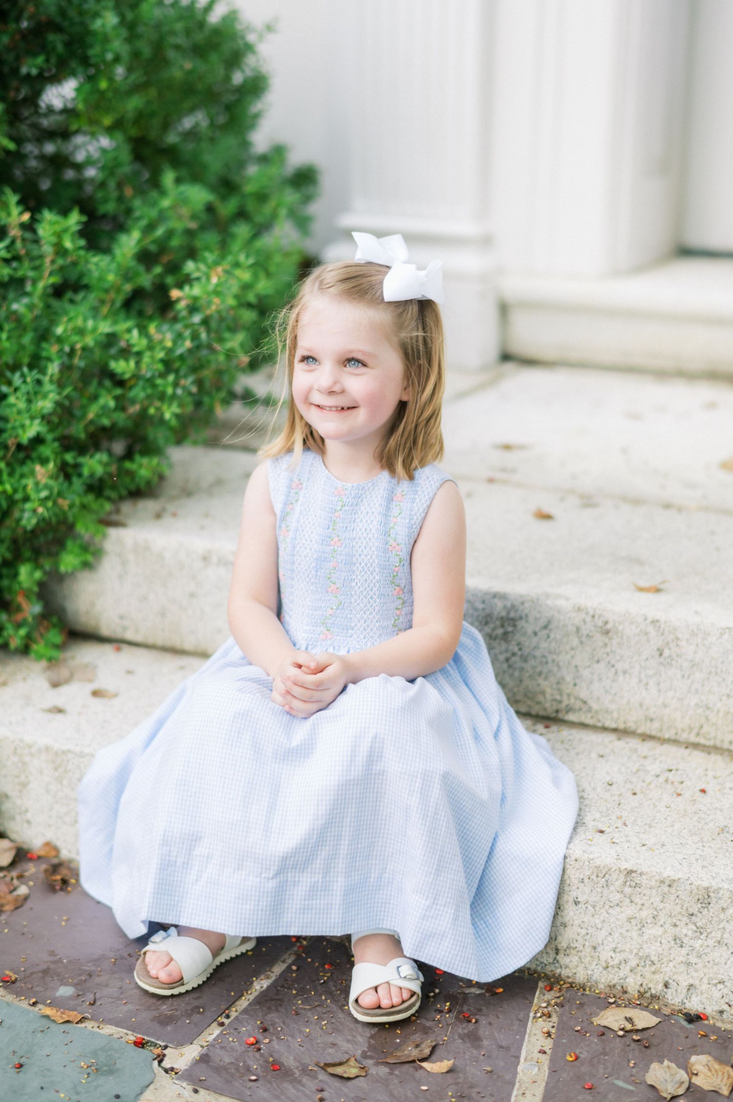 Young child in light blue dress and white bow sitting on stone steps next to greenery.