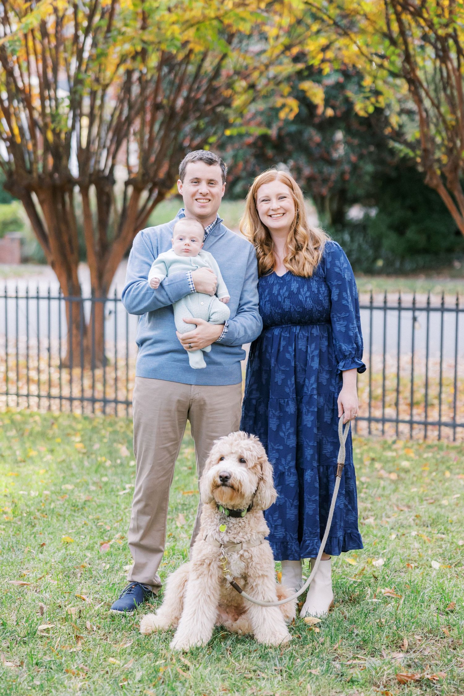 A couple stands with their baby and goldendoodle dog in an autumn garden setting with yellow trees and iron fence.