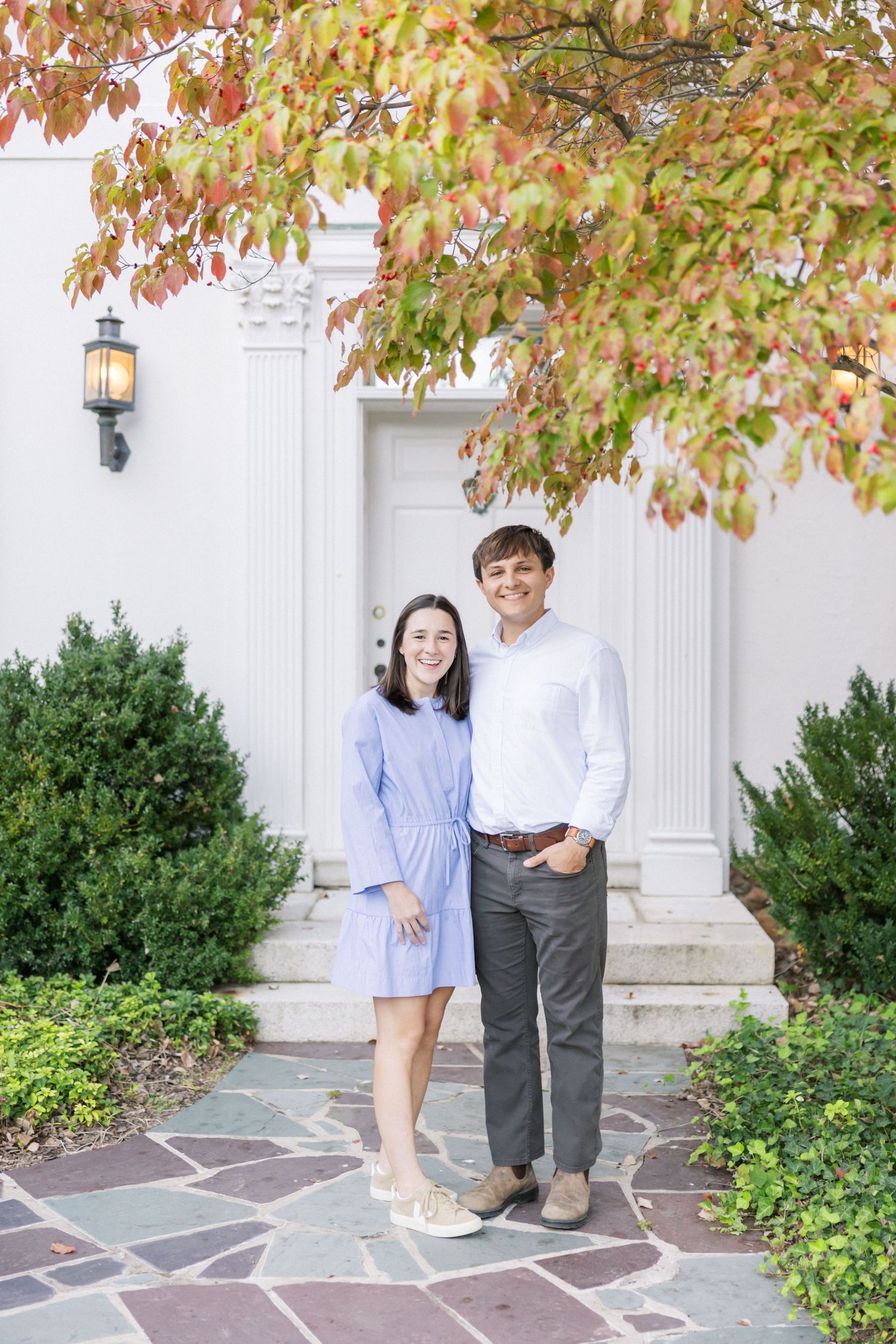 A couple stands on a stone pathway in front of a white doorway, with autumn leaves overhead and shrubs along the sides.