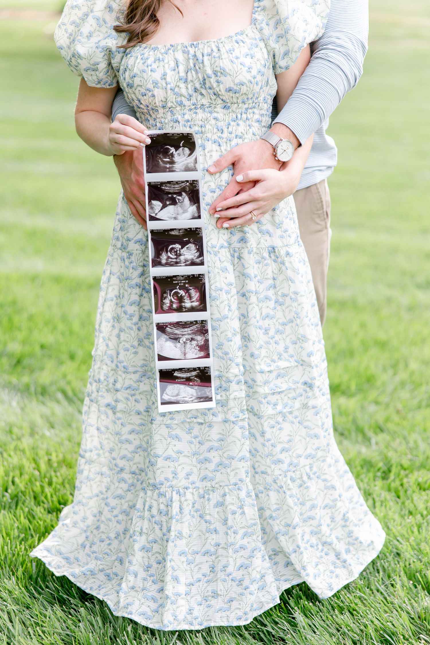 Couple holding baby ultrasound strip while standing in grass, woman wearing floral blue and white maxi dress.