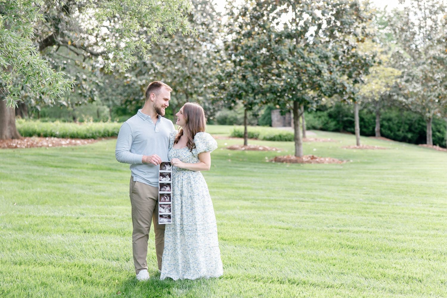 A couple stands together on a lush green lawn surrounded by blooming trees in a tranquil garden setting.