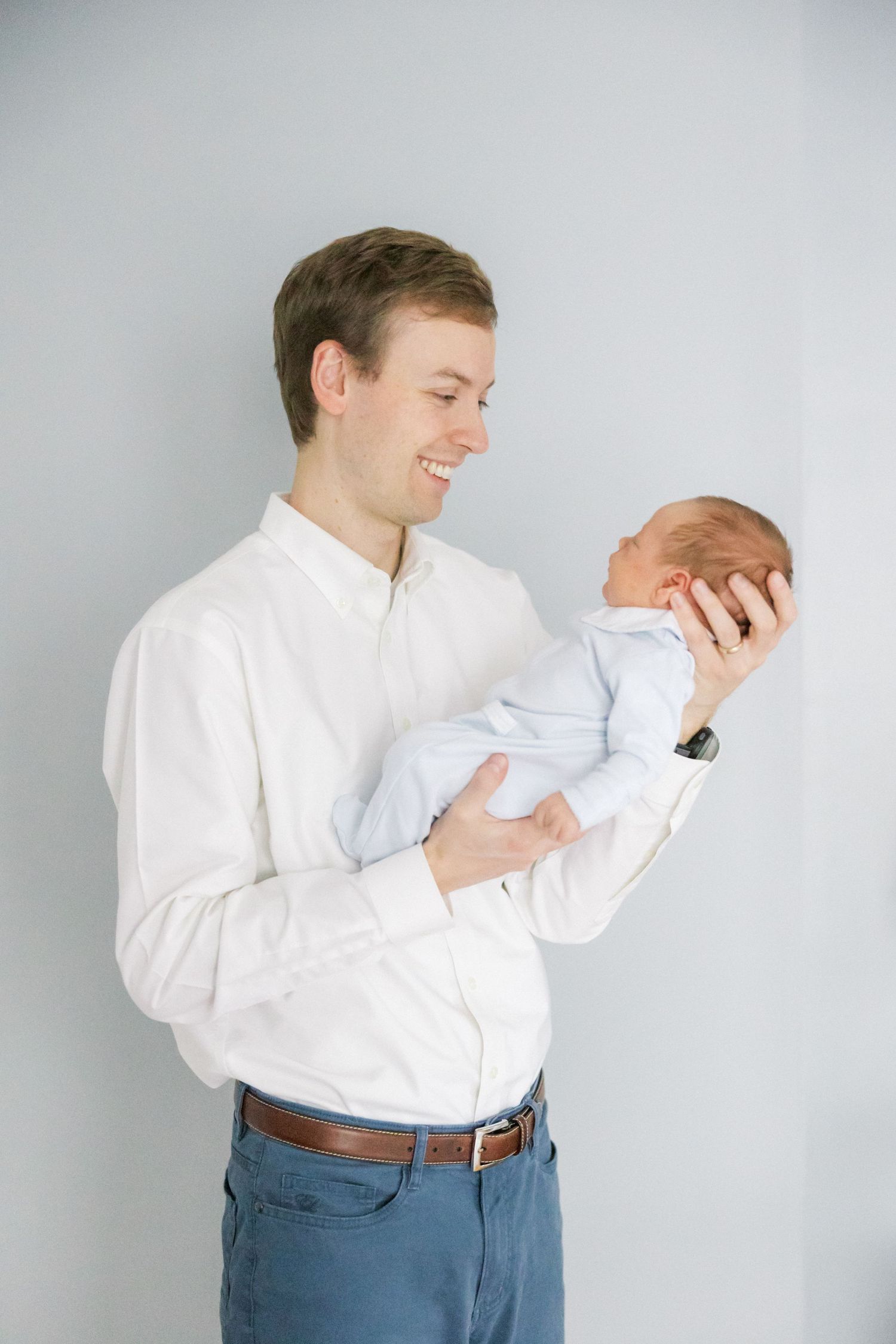 A person in a white shirt and jeans tenderly holds a newborn baby while standing against a gray wall.