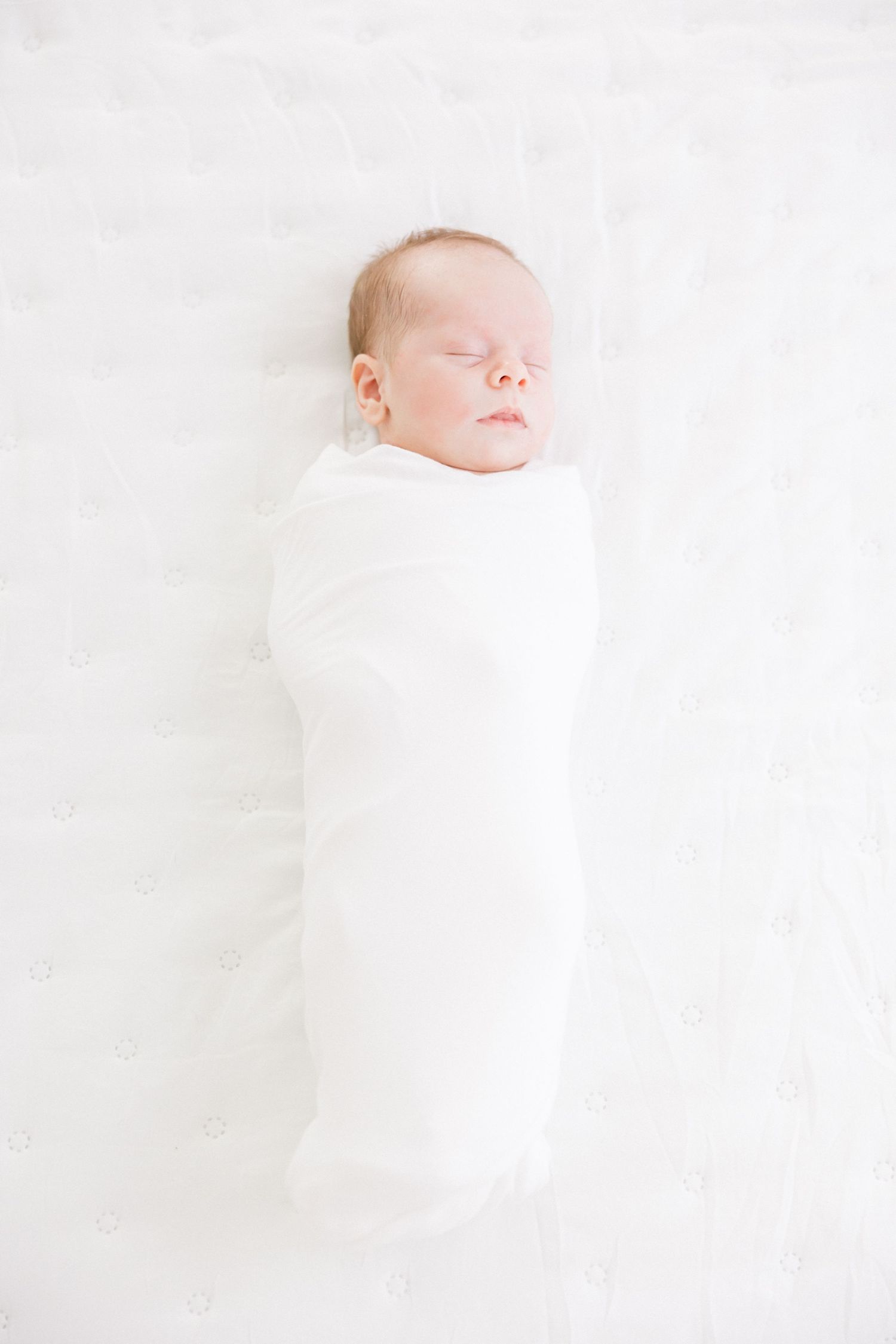 Newborn wrapped snugly in white swaddle blanket on light background during portrait session.