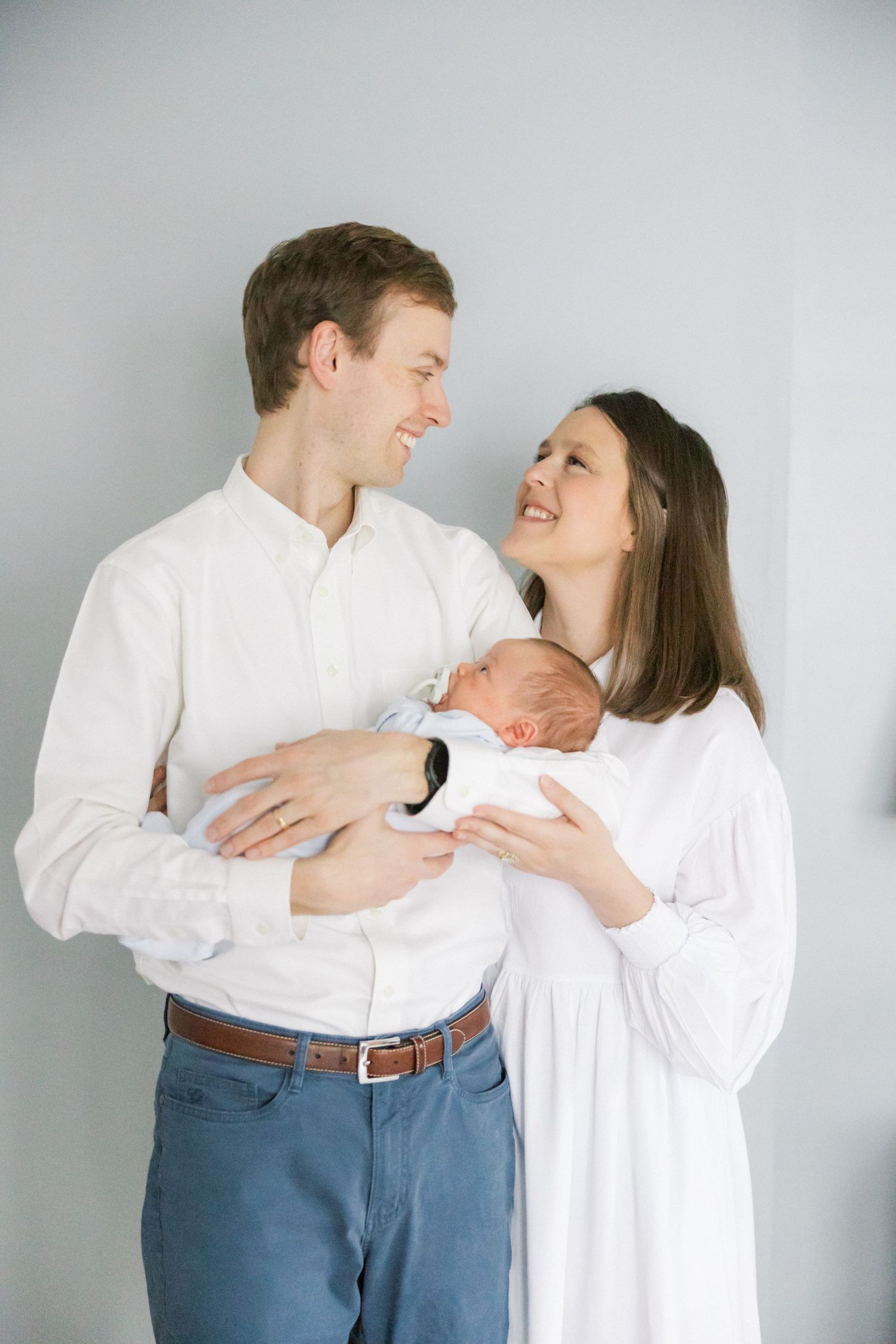 A couple in white shirts share a tender moment while holding their newborn baby against a gray background.