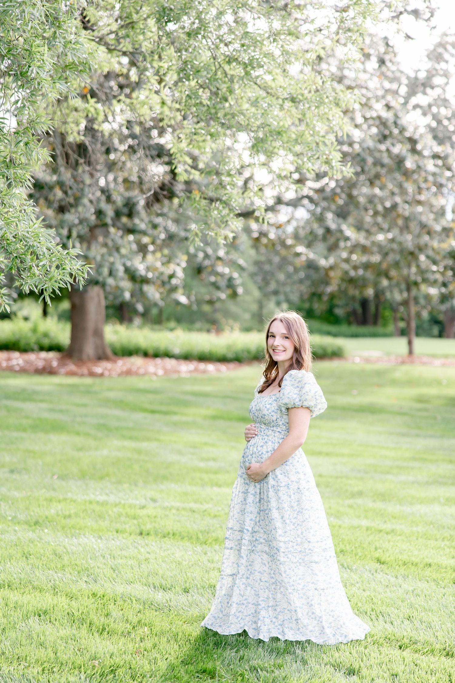 A person in a flowing white floral dress stands in a sunlit garden with blooming trees in springtime.