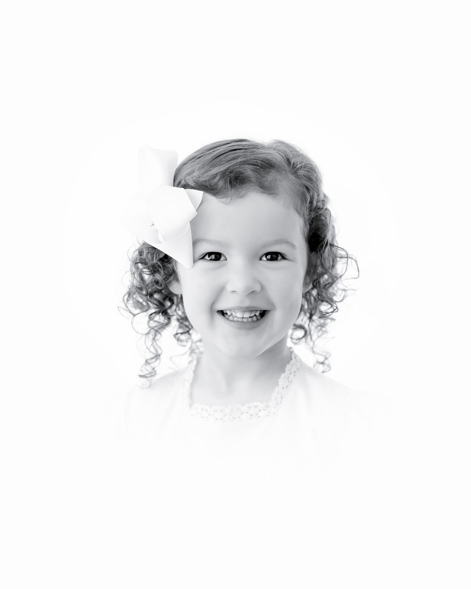 High-key black and white portrait of a laughing toddler with curly hair and white bow.