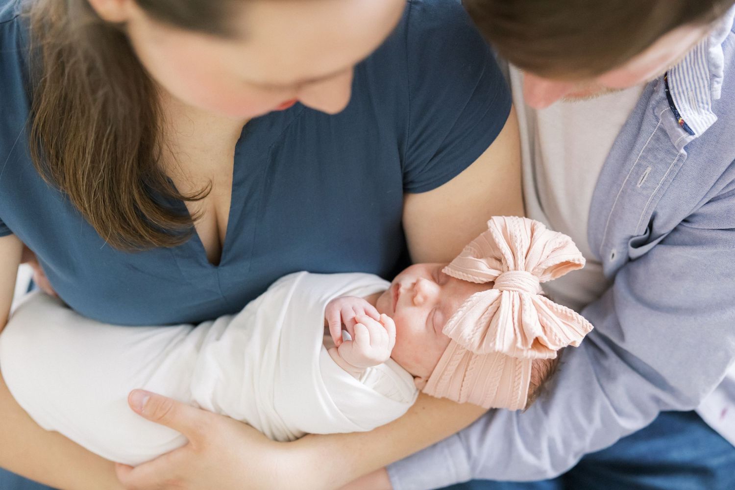 Parents in casual attire admiring their sleeping newborn wearing a pink bow.