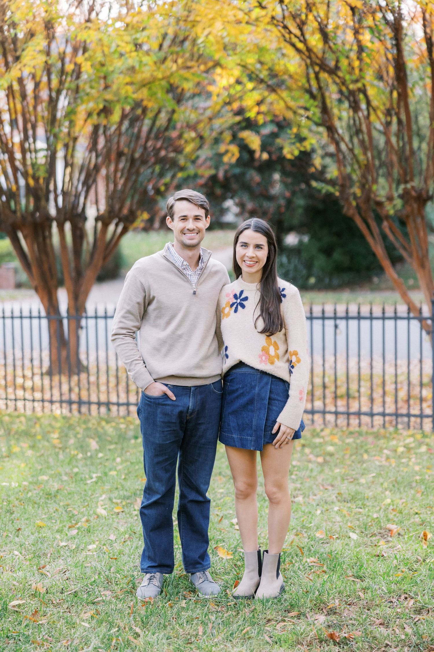 A couple in casual fall attire posing outdoors with autumn foliage and a black iron fence in the background.