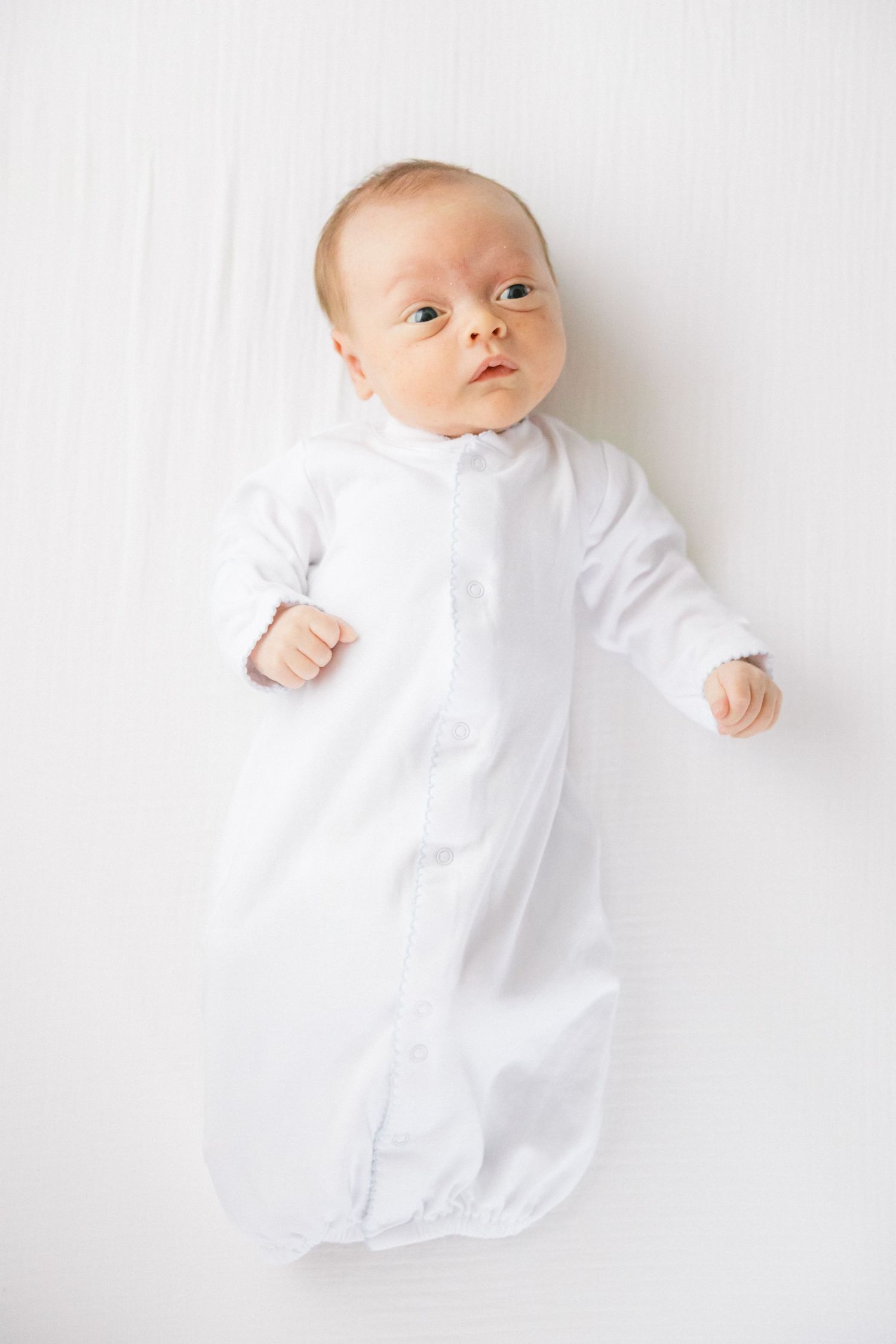 A baby wearing a white gown lies on white fabric looking up at the camera.