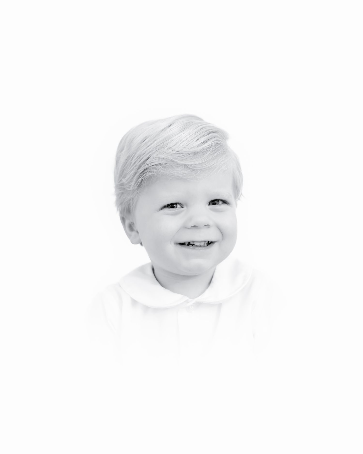 Black and white portrait of a young child with light hair smiling warmly against a white background.