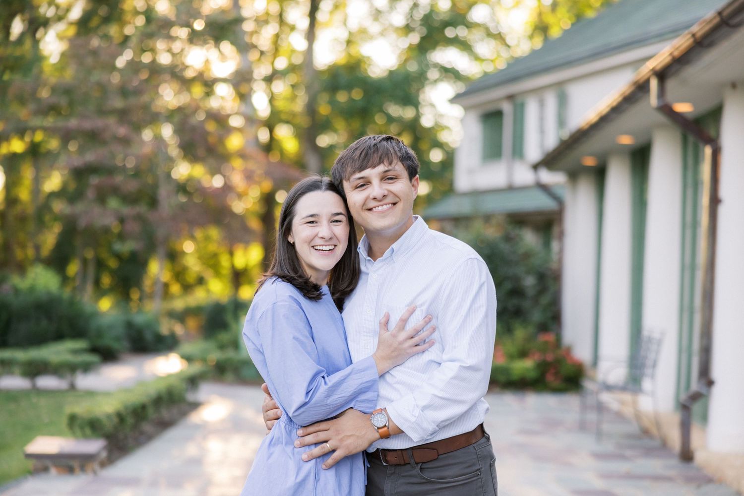 A couple smiling and embracing outdoors with a house and garden behind them.