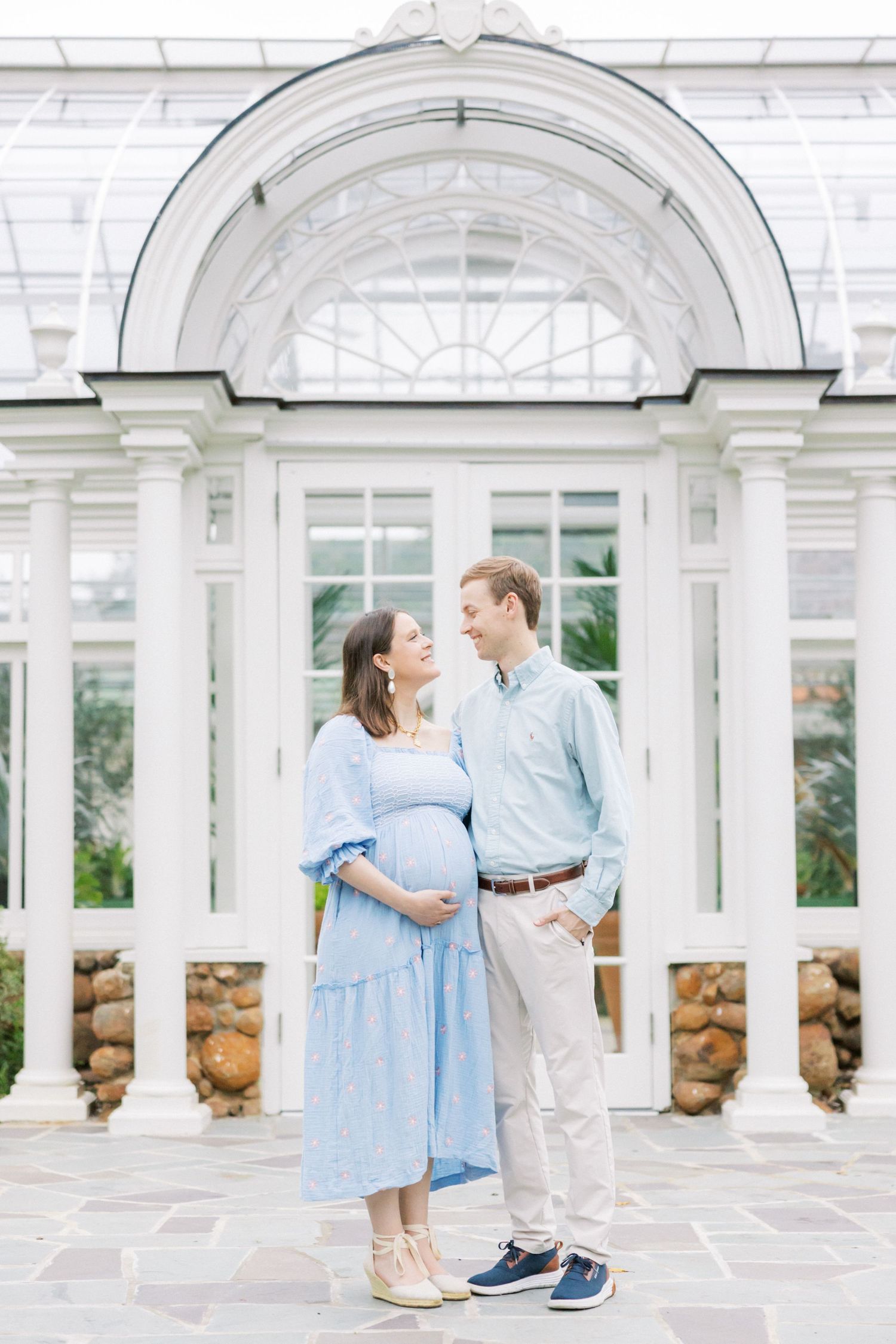 Couple poses under white architectural arch during maternity photoshoot.