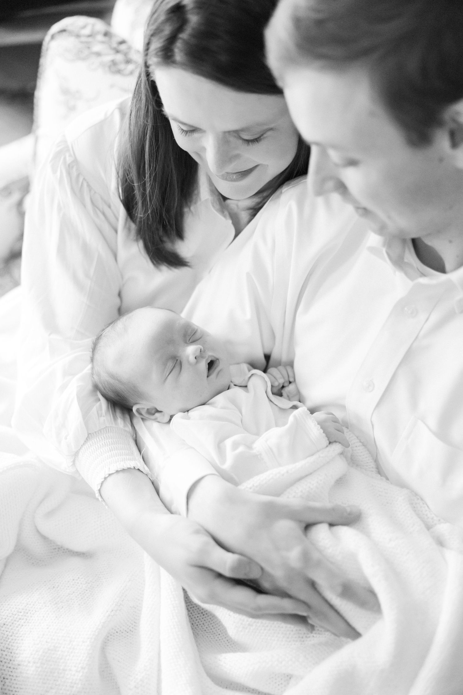Black and white portrait of parents gazing at their newborn baby in white clothing.