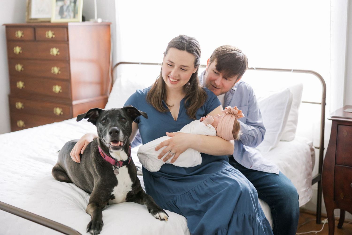 A couple sits on bed with their black dog while holding a newborn baby.