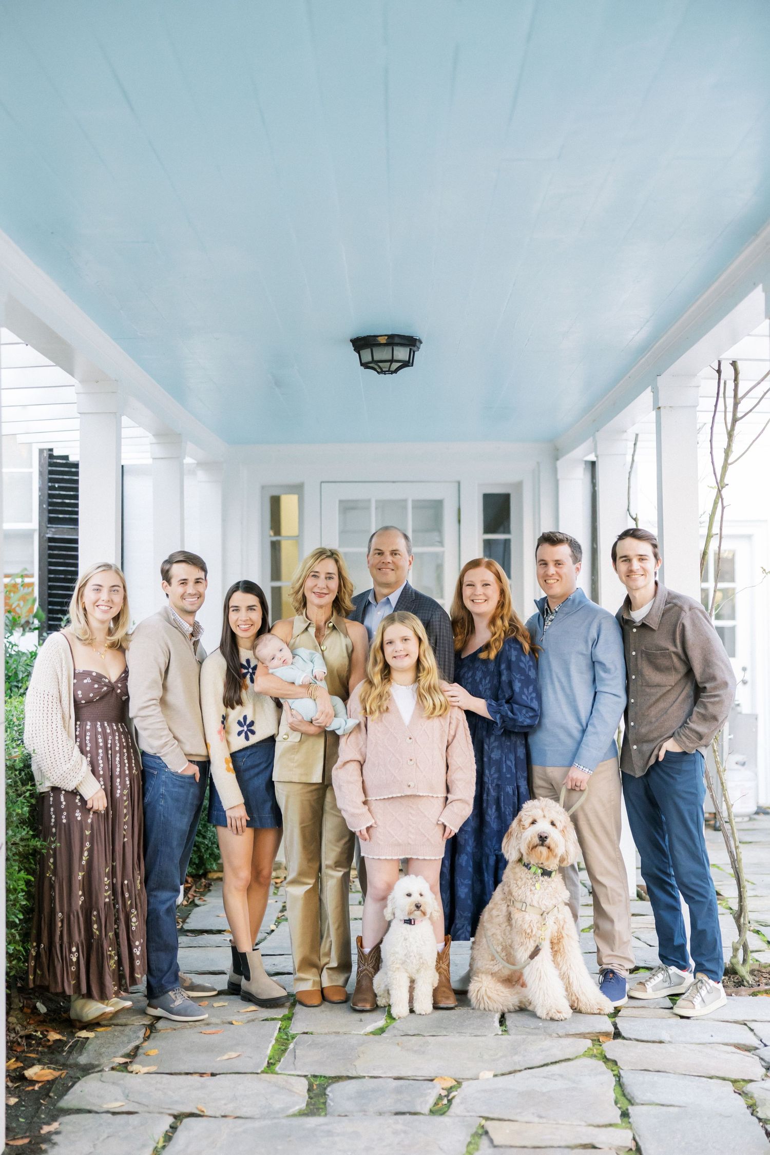 Extended family portrait on a white porch with two dogs and a baby in coordinated neutral outfits.