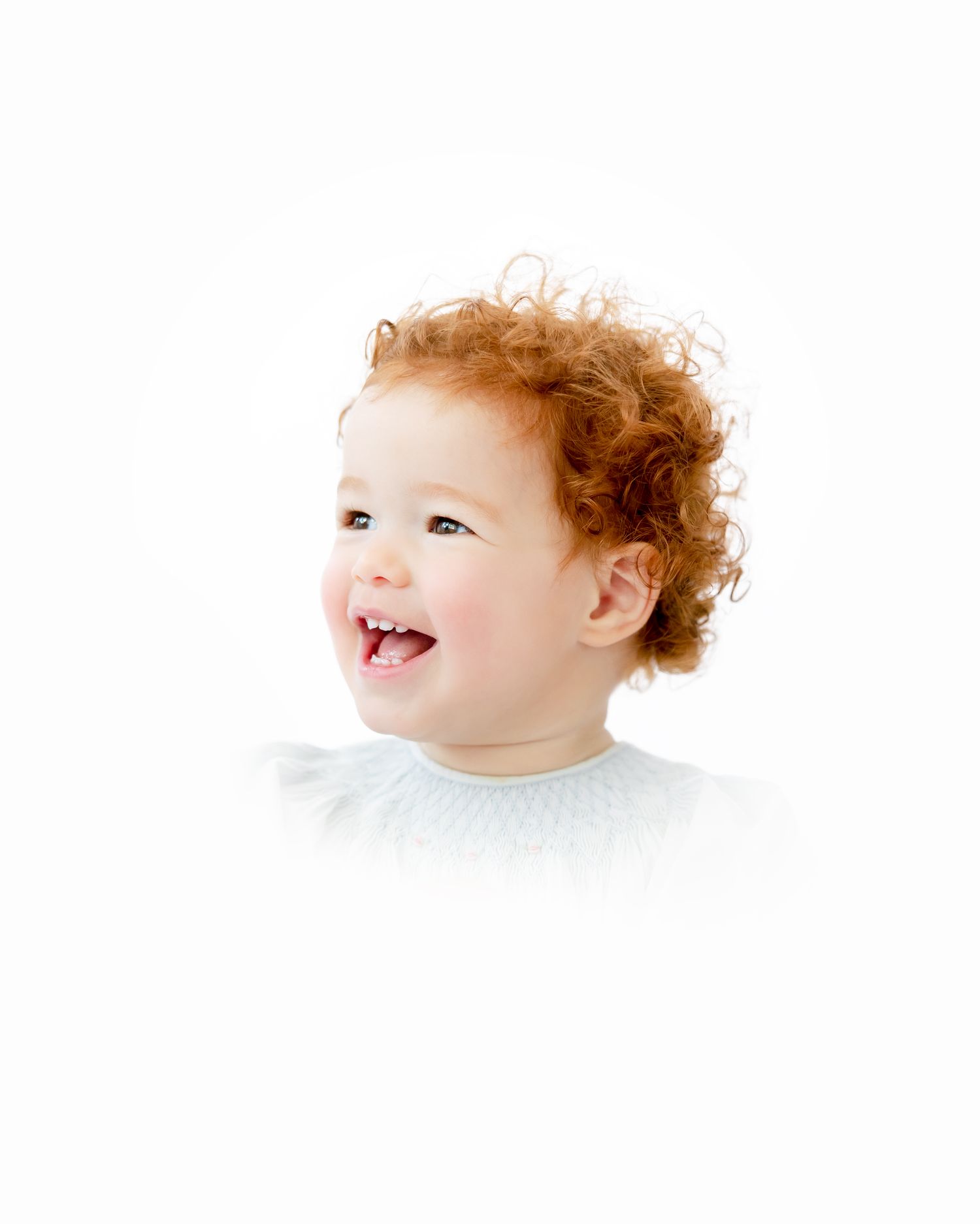 A smiling toddler with auburn curly hair wearing a white top against a white background.