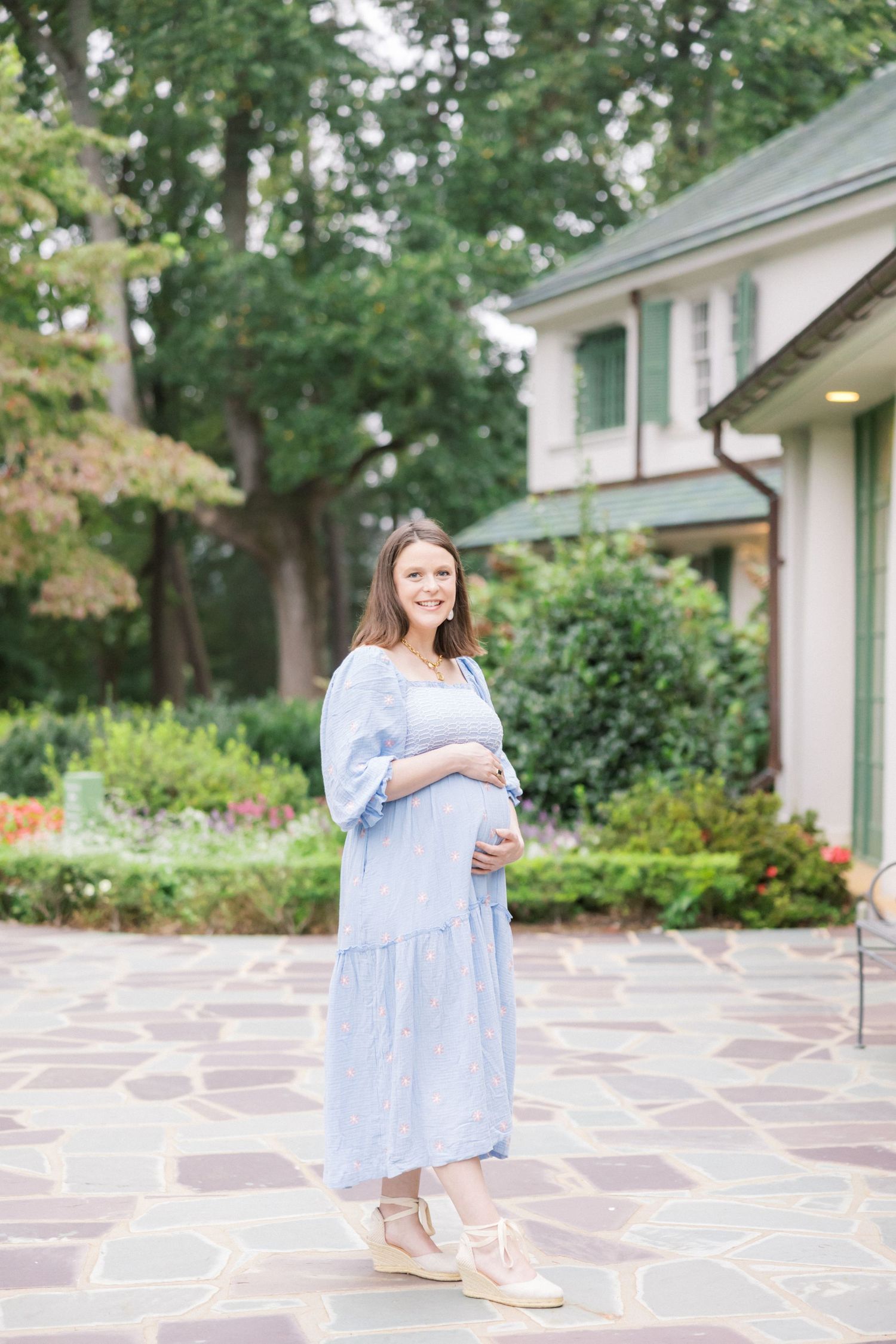 A woman in a flowing light blue dress stands on a stone patio outside an elegant house with gardens.