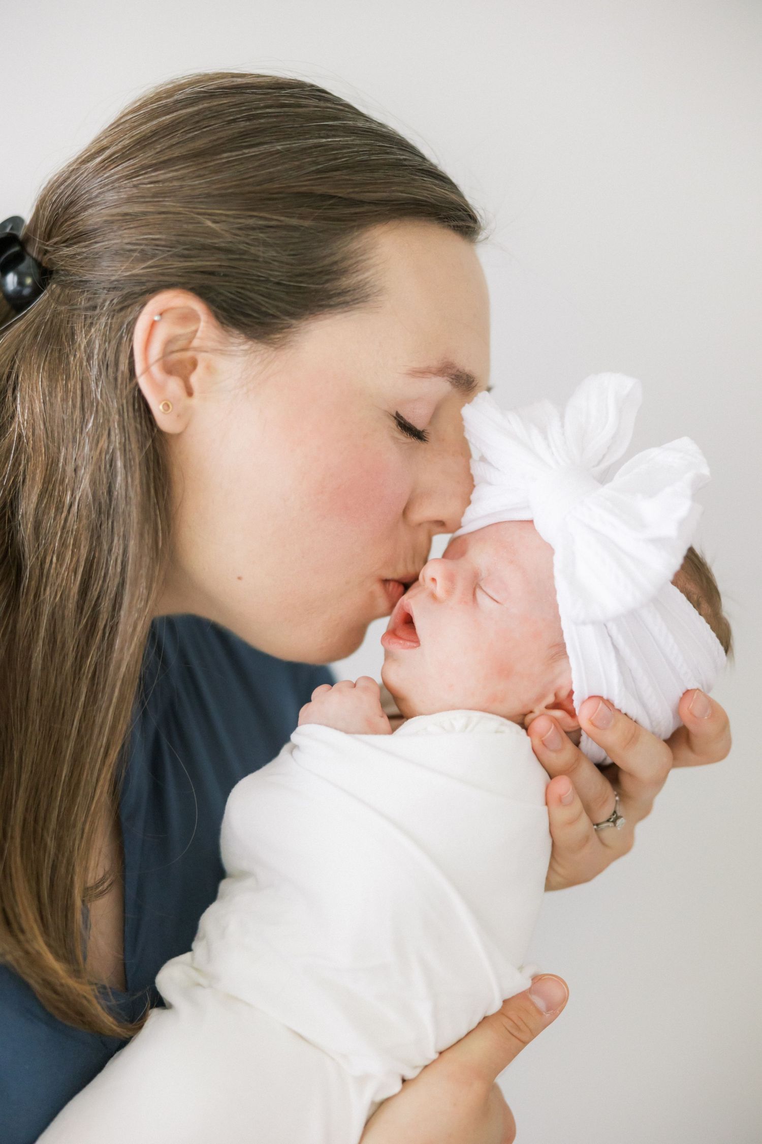 Tender moment between caregiver and infant wrapped in white with bow during portrait session.