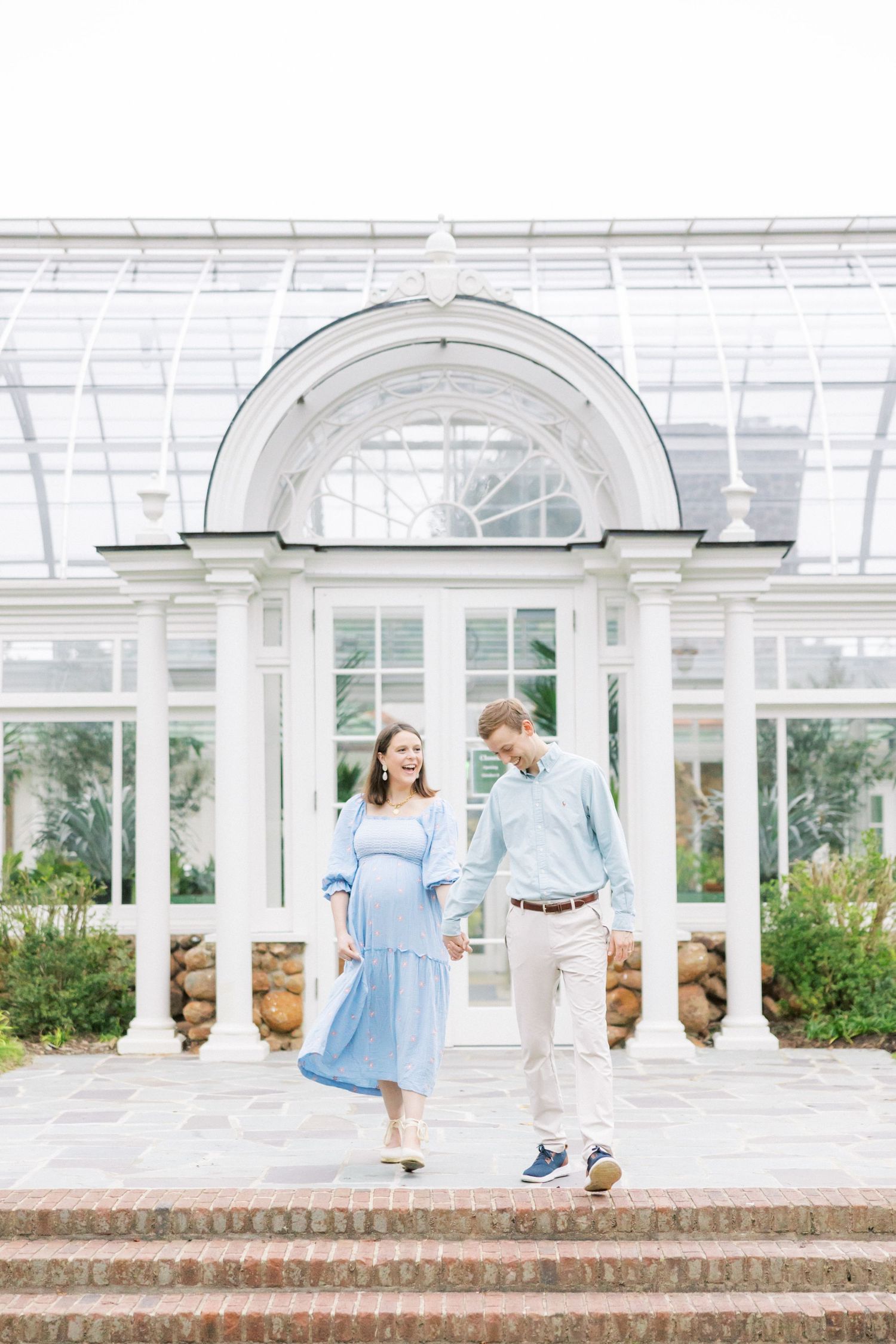 Expecting couple stands on steps of white conservatory building during photoshoot.