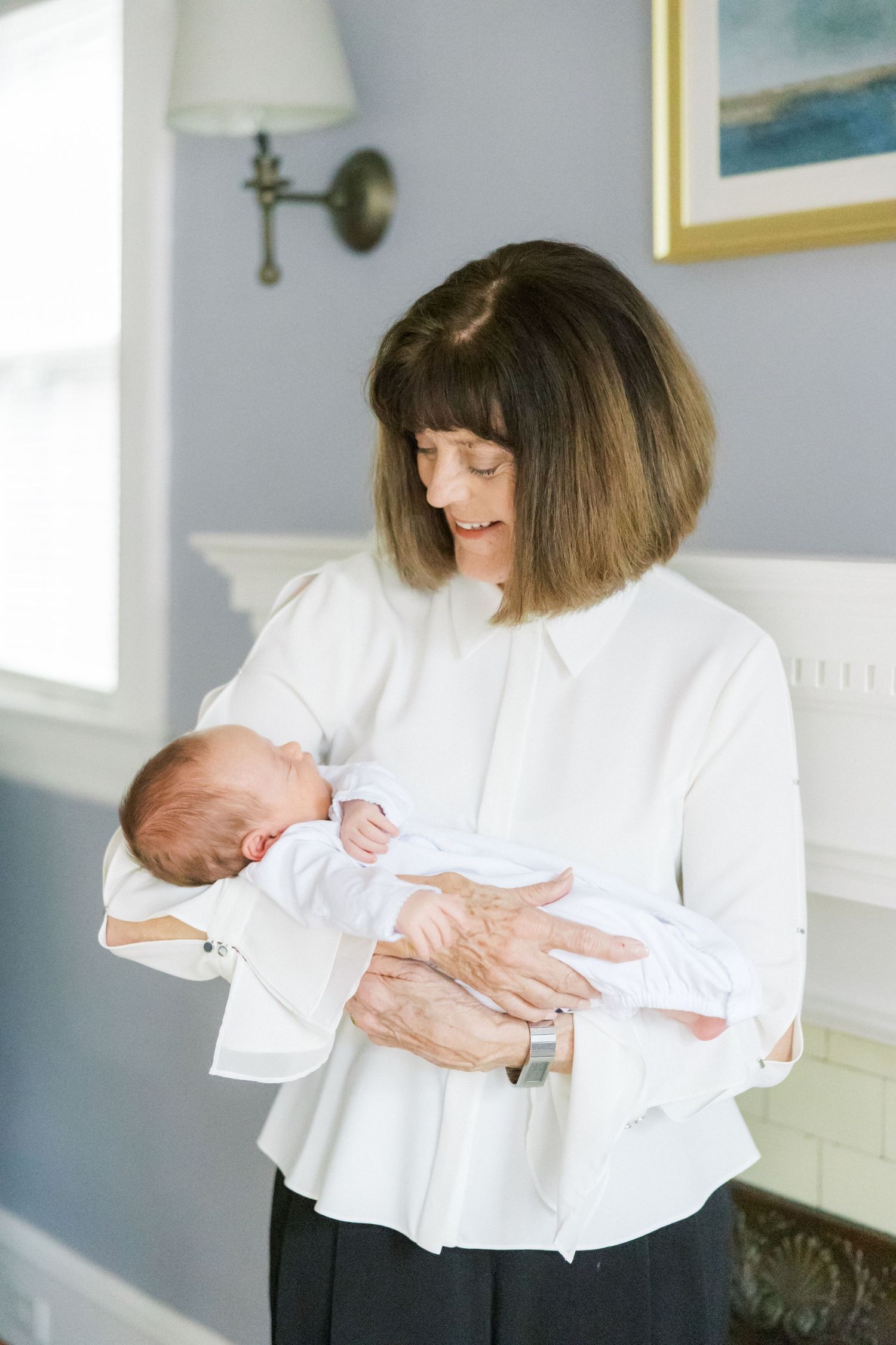 Woman in white blouse carefully holds sleeping newborn in sunny room with blue walls.