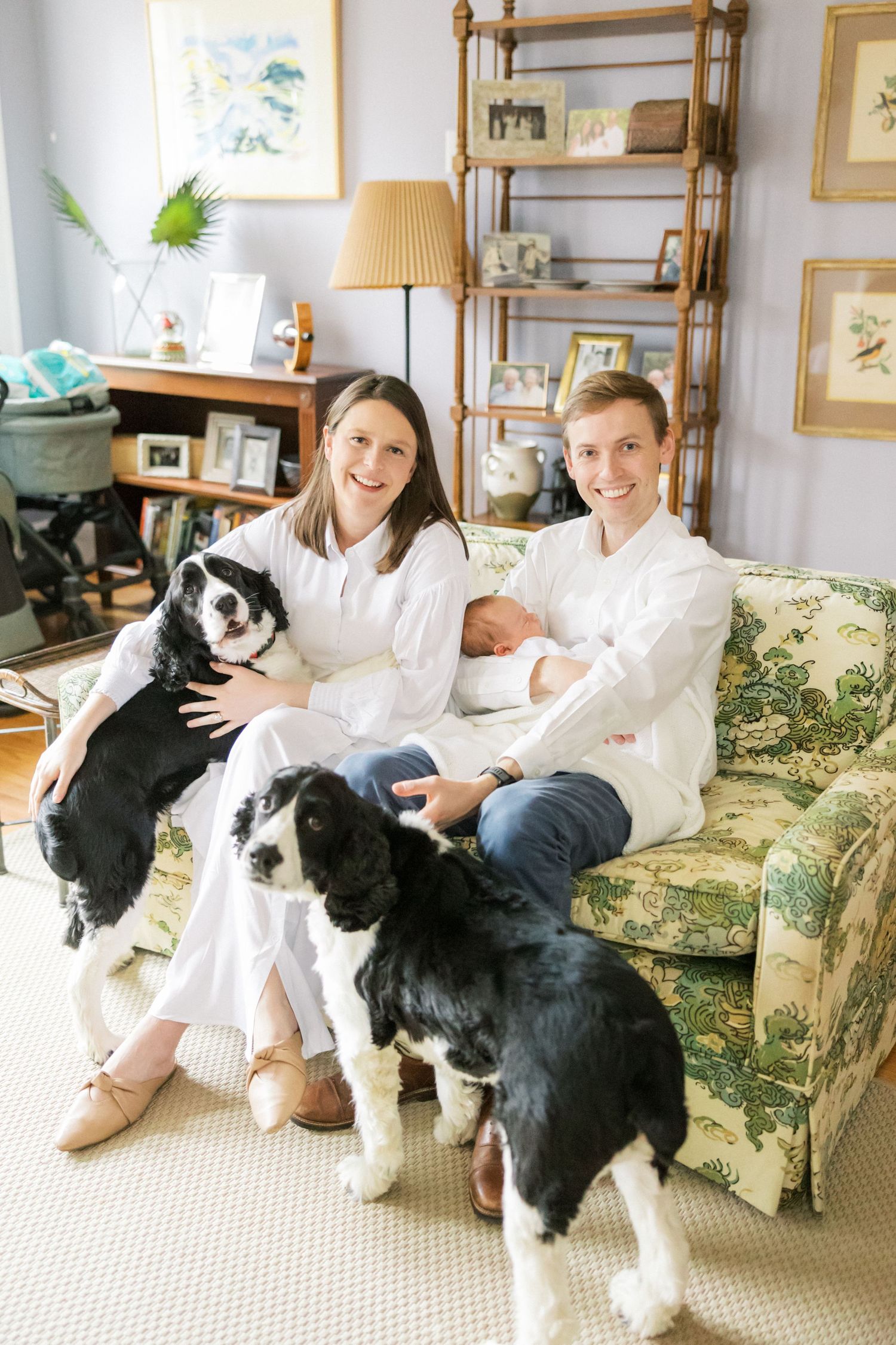 Family poses on floral couch with their newborn baby and two black and white dogs.