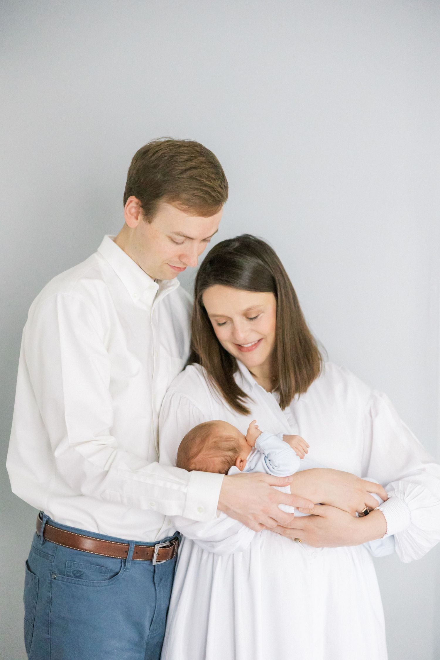 A couple in white shirts tenderly holds their newborn baby against a gray backdrop.