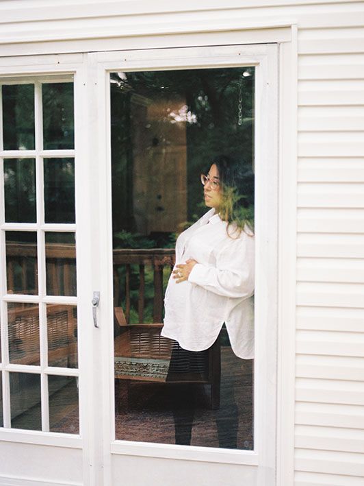 A figure stands at glass doors in white attire against the backdrop of a white clapboard house exterior.