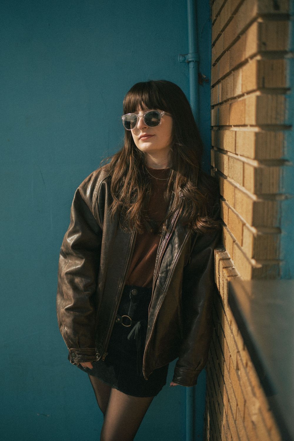 A stylish figure in sunglasses and brown leather jacket poses against turquoise wall and brick in moody lighting.