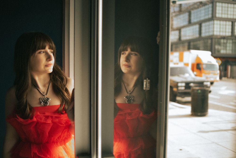 A reflection in a window shows a person wearing a red dress against an urban street backdrop.