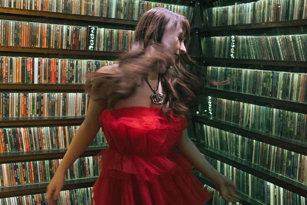 A person in a red strapless dress poses playfully against a wall of bookshelves filled with colorful books and media.