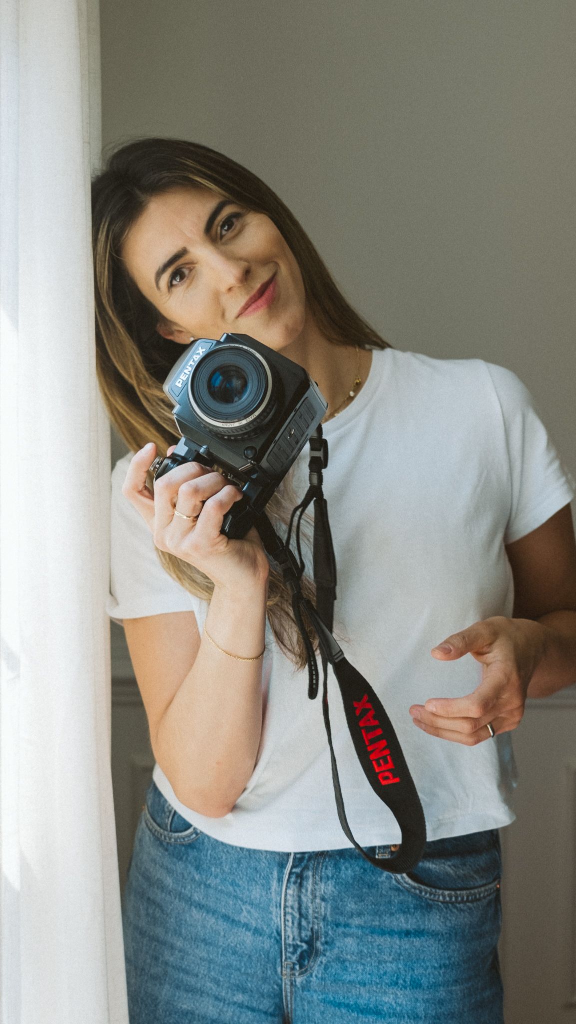 A photographer in a white t-shirt and jeans poses with a professional DSLR camera against a light colored wall.