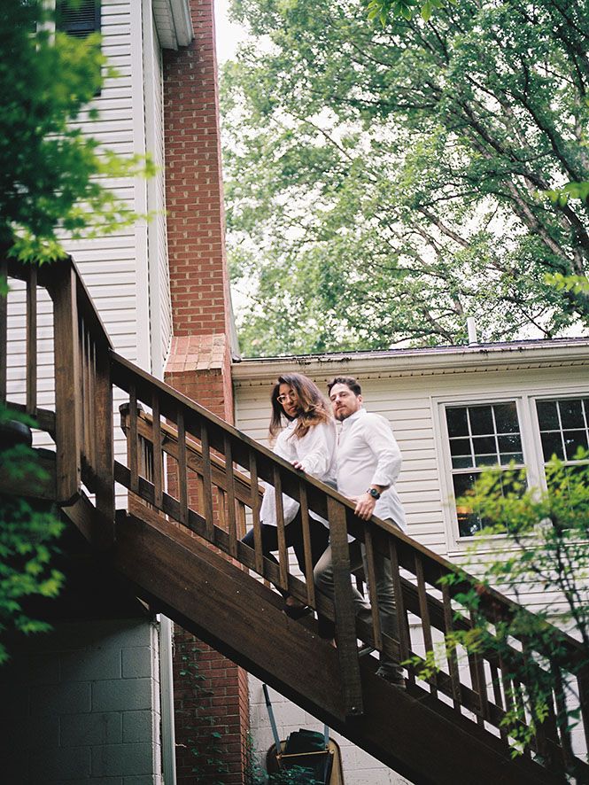 A couple descends a wooden outdoor staircase at a white residential house with trees blooming in the background.