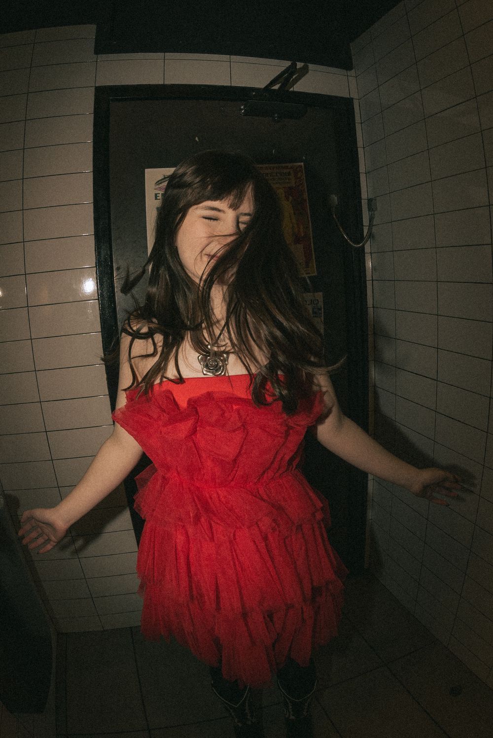 Someone wearing a red ruffled dress dances joyfully in a dimly lit hallway with tiled walls.