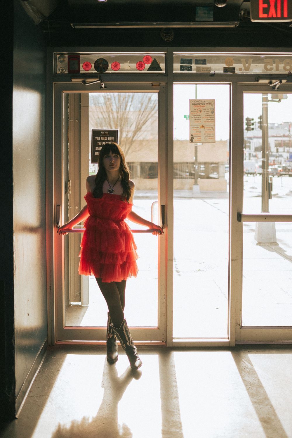 A silhouetted figure in a flowy red dress poses against a bright doorway entrance, casting dramatic shadows on the floor.