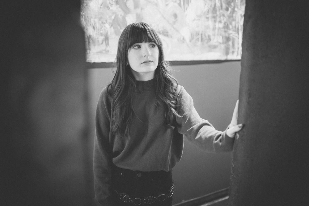 Black and white portrait of someone in a turtleneck sweater standing near a window with natural light streaming in.