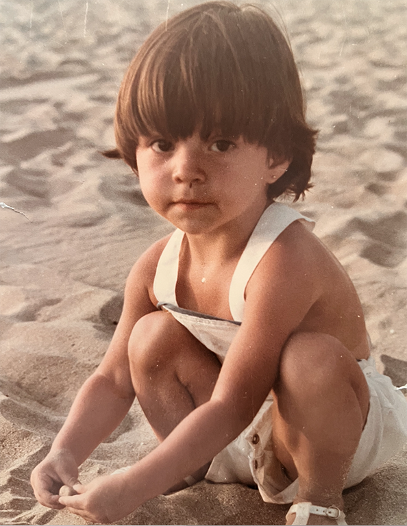 Small child with brown hair sitting and playing in sand on a beach, wearing a white tank top.