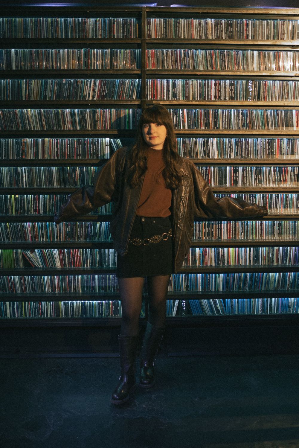 Someone in a leather jacket and skirt poses against a wall of vinyl records in a dimly lit record store.