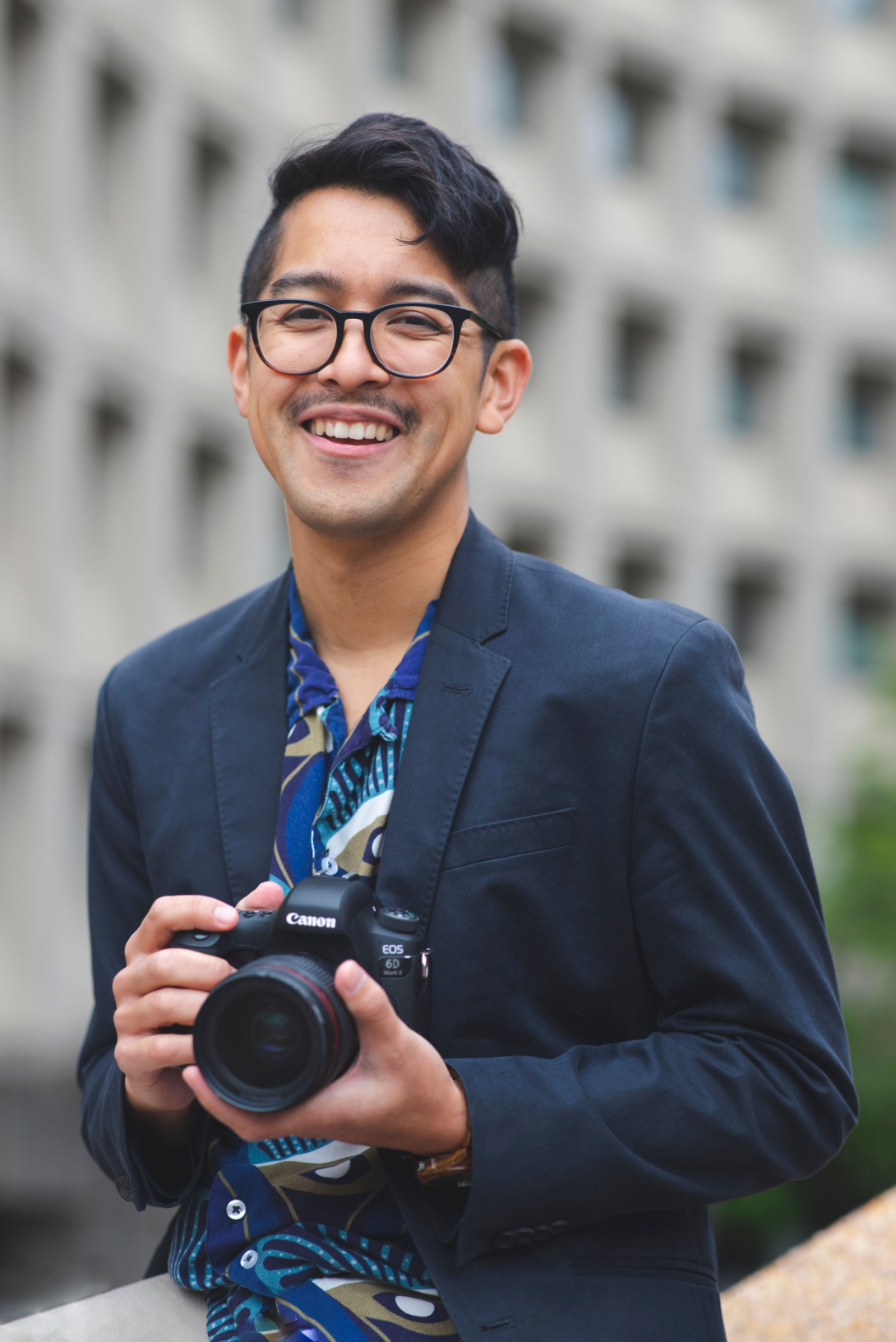 Professional photographer in navy blazer holding camera outdoors with joyful expression.